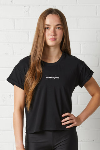 Young woman wearing a NorthByOne black Stella Boxy Tee with rolled sleeves standing against a white brick wall background