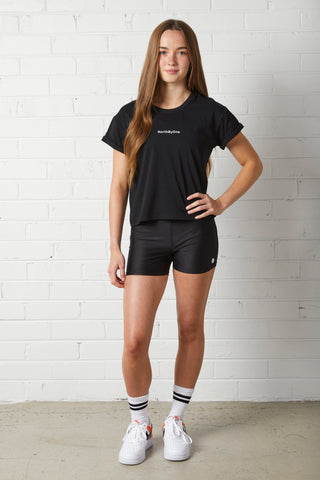 Young woman wearing a NorthByOne black Stella Boxy Tee with cuffed sleeves and shorts standing against a white brick wall