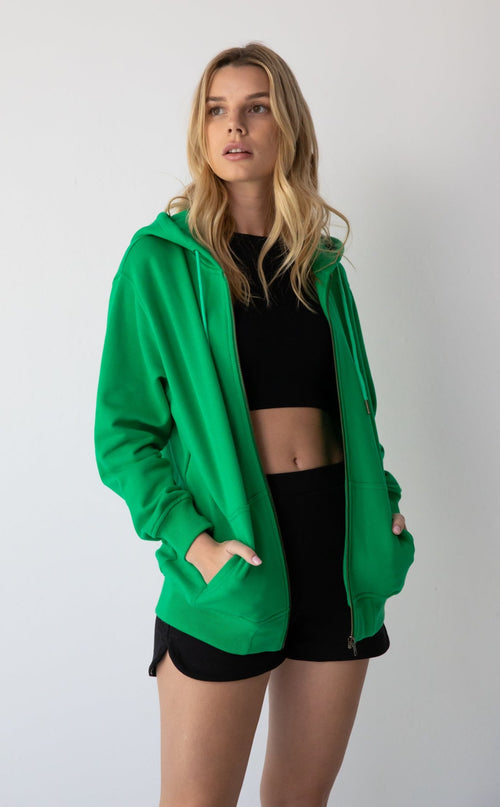 Young woman wearing a NorthByOne Olive Zipper Hoodie in Emerald over black crop top and shorts against a plain white background