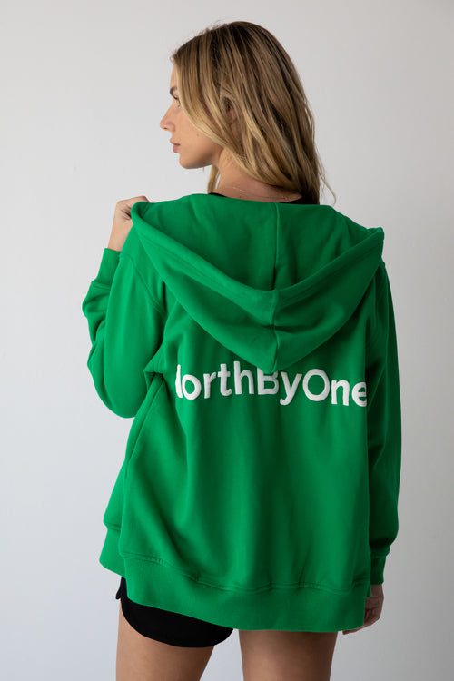 Woman wearing an Olive Zipper Hoodie in Emerald with NorthByOne logo on the back against a plain white background