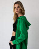 Young woman wearing a NorthByOne Olive Zipper Hoodie in Emerald with black crop top and shorts standing against a plain background