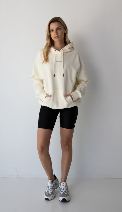 Woman wearing a NorthByOne Frankie Hoodie in vanilla with black biker shorts and sneakers standing against a plain white background