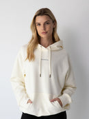 Young woman wearing a NorthByOne Frankie Hoodie in vanilla with a hood and logo, standing against a plain white background