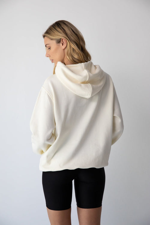 Young woman wearing a NorthByOne Frankie Hoodie in vanilla with hood up and black shorts standing against a plain background