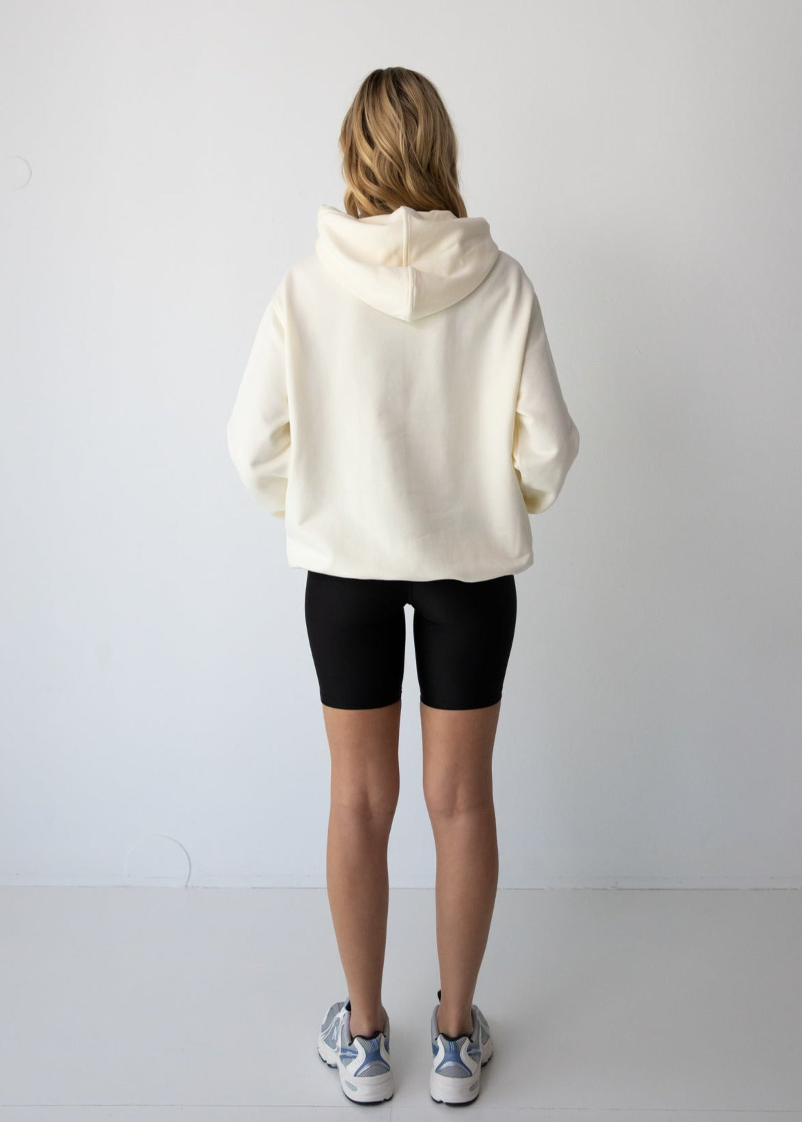 Woman wearing a NorthByOne Frankie Hoodie in vanilla paired with black biker shorts and sneakers against a plain white background