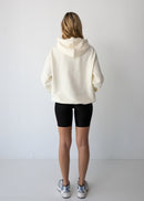 Woman wearing a NorthByOne Frankie Hoodie in vanilla paired with black biker shorts and sneakers against a plain white background