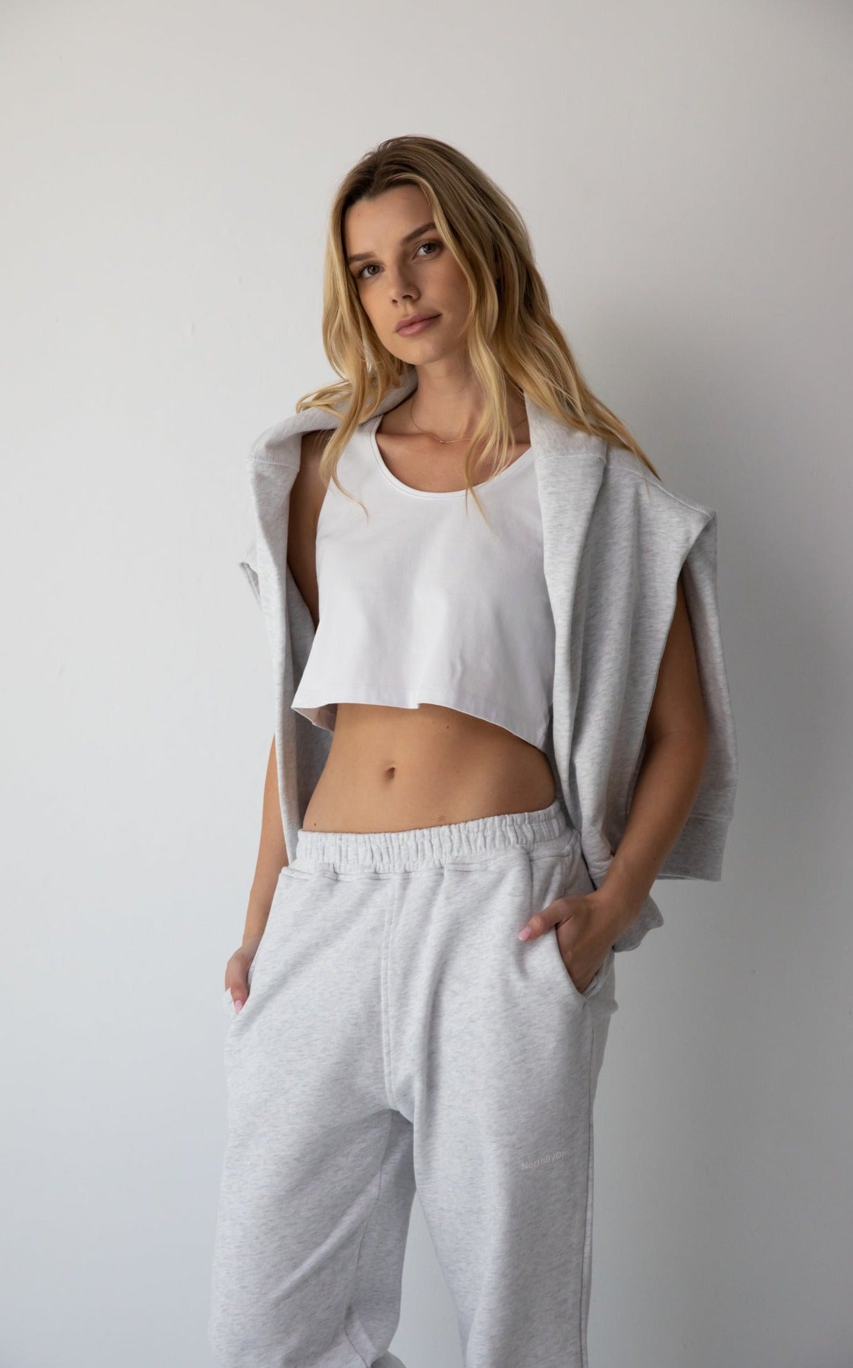 Woman wearing the NorthByOne Emma Baggy Sweat Pant in Marle Grey, with a white cropped tank top and sleeveless jacket against plain background