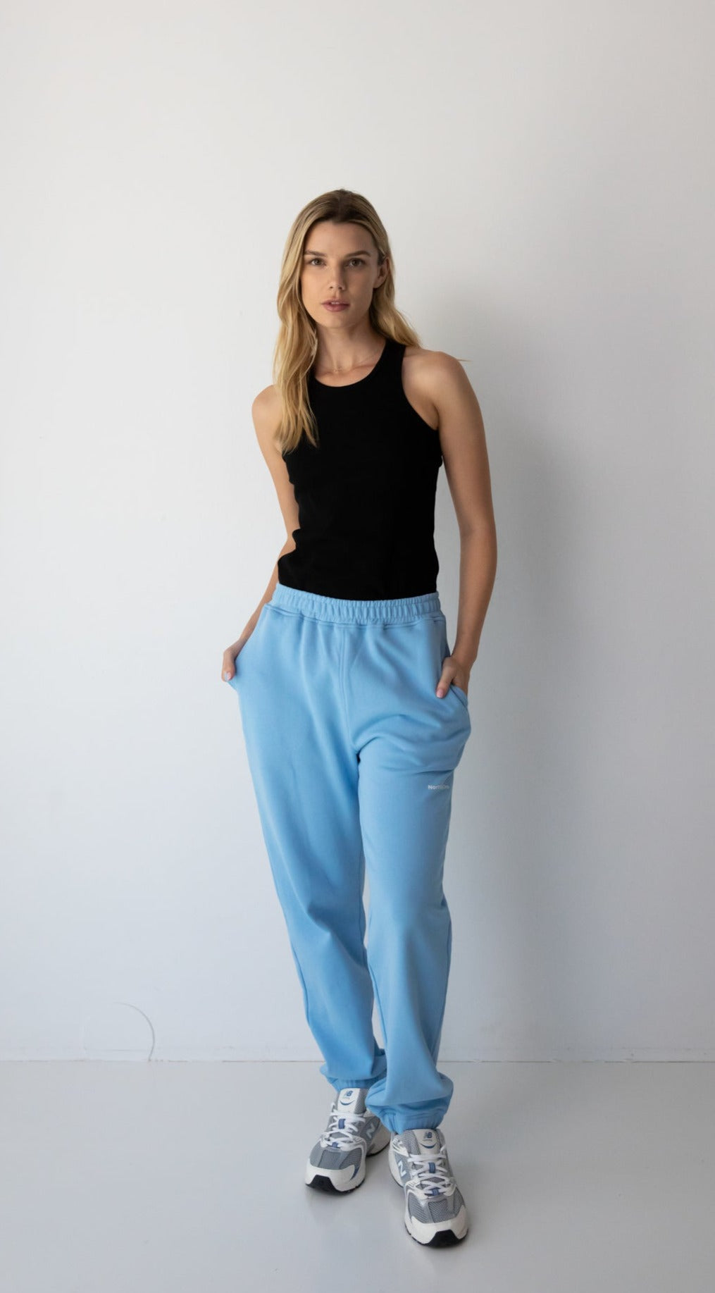 Woman wearing the NorthByOne Emma Baggy Sweat Pant in Colbalt Blue, with a black sleeveless top and sneakers standing against a plain white background