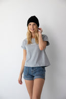 Smiling woman wearing a black beanie, gray NorthByOne t-shirt, and Charlotte Denim Short standing against a white background