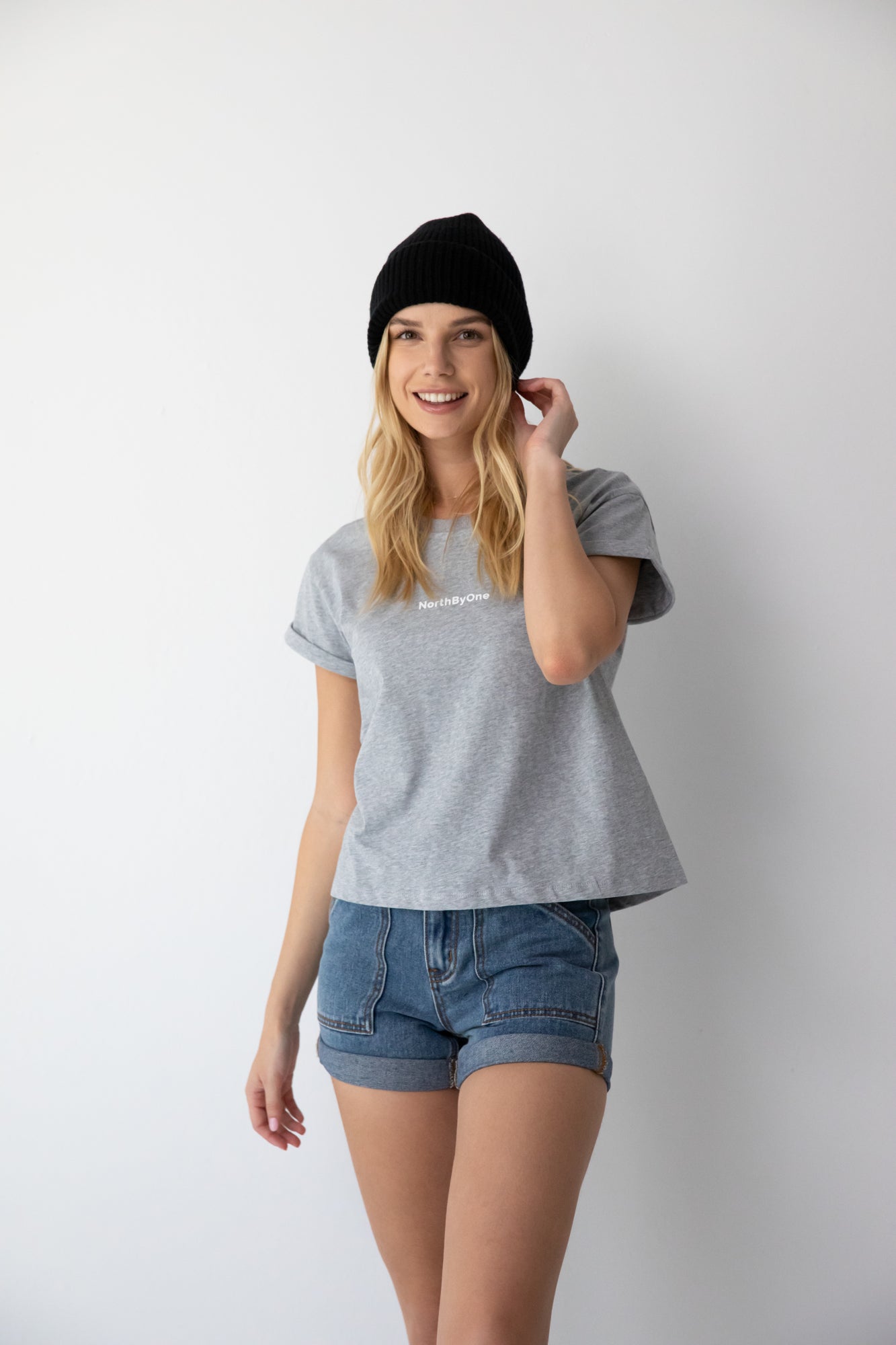Smiling woman wearing a black beanie, gray NorthByOne t-shirt, and Charlotte Denim Short standing against a white background