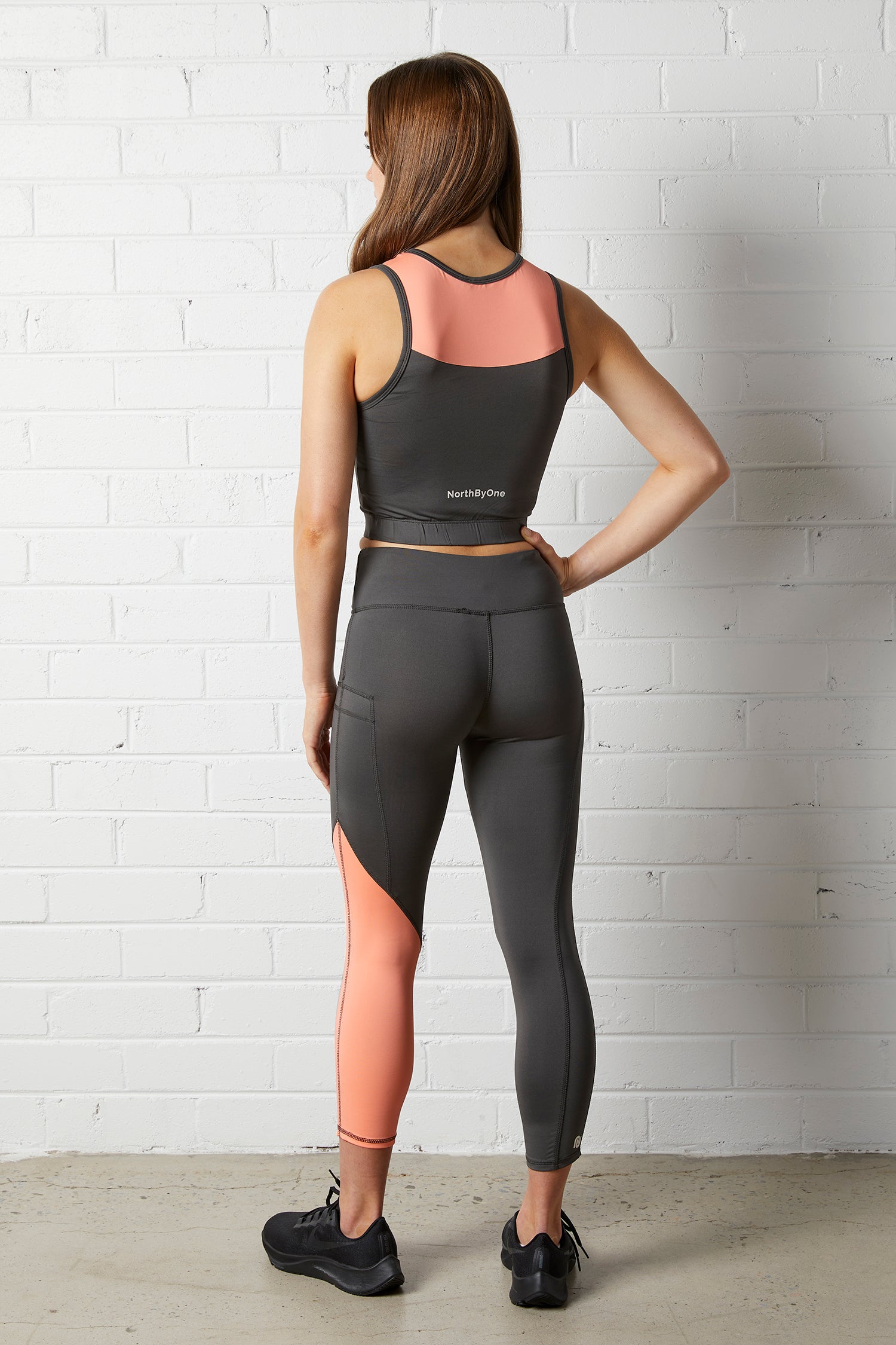 Woman wearing the NorthByOne Cara Three Quarter Tight - Slate Grey / Neon Pink with matching top standing against a white brick wall