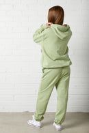 Woman wearing the NorthByOne Emma Baggy Sweat Pant in Avocado, and matching hoodie standing against a white brick wall from the back