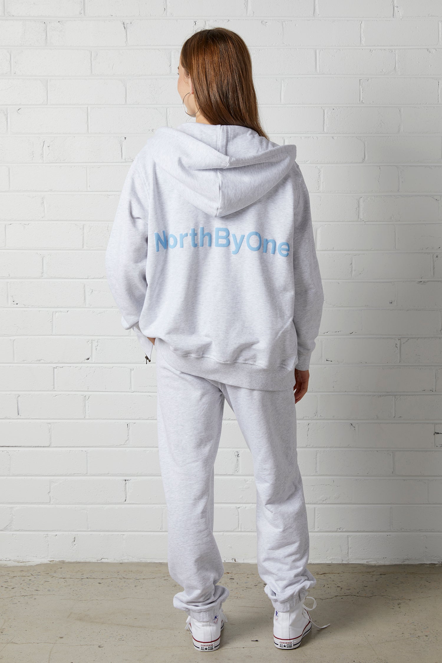 Woman wearing a NorthByOne Olive Zipper Hoodie in Marle Grey with matching pants standing against white brick wall