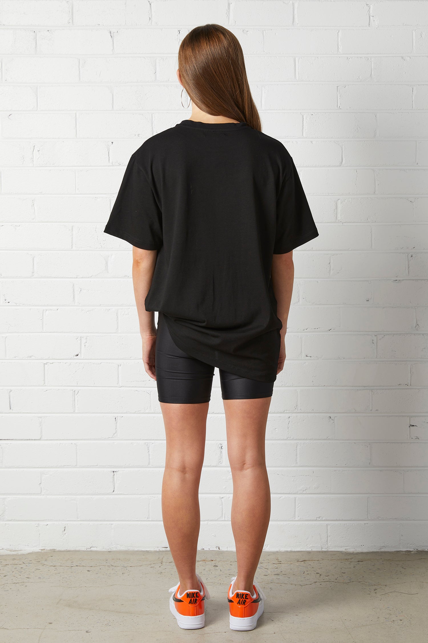 Young woman wearing Lucy Oversized Tee | NorthByOne in black with black bike shorts and orange sneakers against white brick wall