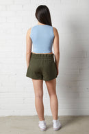 Woman wearing a NorthByOne Aria Paperbag Short in a Olive colour, paired with a light blue sleeveless top standing against a white brick wall