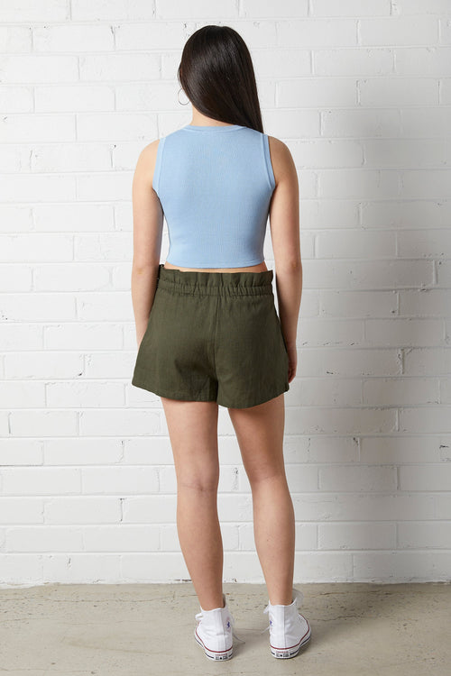 Woman wearing a NorthByOne Aria Paperbag Short in a Olive colour, paired with a light blue sleeveless top standing against a white brick wall