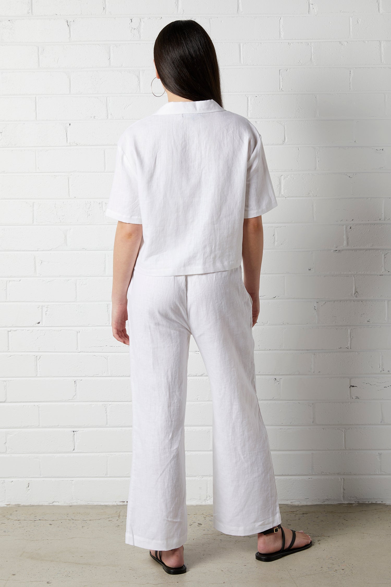 Woman with long dark hair wearing NorthByOne Sadie Linen Pant in white and a matching top standing against a white brick wall