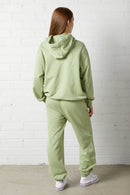 Woman wearing the NorthByOne Frankie Hoodie in avocado and matching pants standing with back to camera against white brick wall