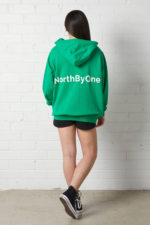 Woman wearing an oversized NorthByOne Olive Zipper Hoodie in Emerald with NorthByOne logo on back standing against white brick wall