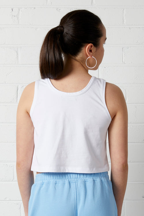 The back of a woman wearing a NorthByOne Allegra Crop singlet in a white colour with a blue pair of sweats, looking over her shoulder against a white brick wall