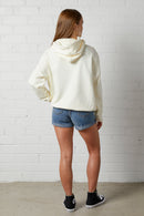 Woman wearing a NorthByOne Frankie Hoodie in vanilla with denim shorts and black sneakers standing against a white brick wall