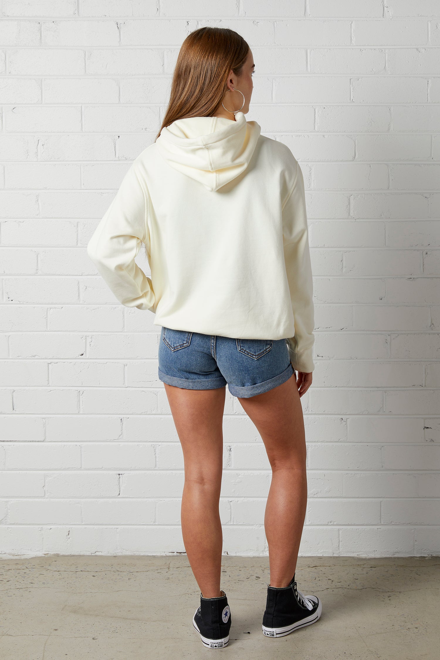 Woman wearing a NorthByOne Frankie Hoodie in vanilla with denim shorts and black sneakers standing against a white brick wall