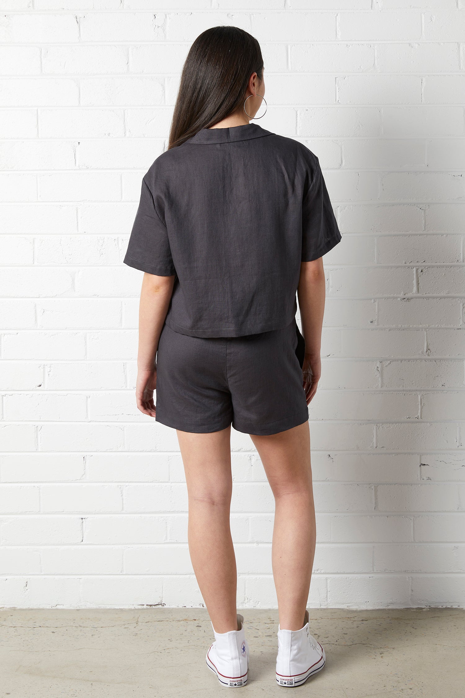 Woman wearing the NorthByOne Zoe Linen Shirt in Charcoal with matching shorts standing against a white brick wall