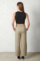 Woman wearing NorthByOne Sadie Linen Pant in Sage Green with elasticated waistband and black sleeveless crop top standing against white brick wall