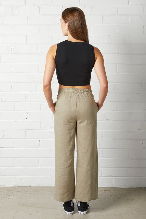 Woman wearing NorthByOne Sadie Linen Pant in Sage Green with elasticated waistband and black sleeveless crop top standing against white brick wall