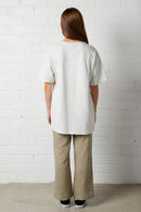 Woman with long brown hair wearing Lucy Oversized Tee | NorthByOne in Marle Grey standing against a white brick wall