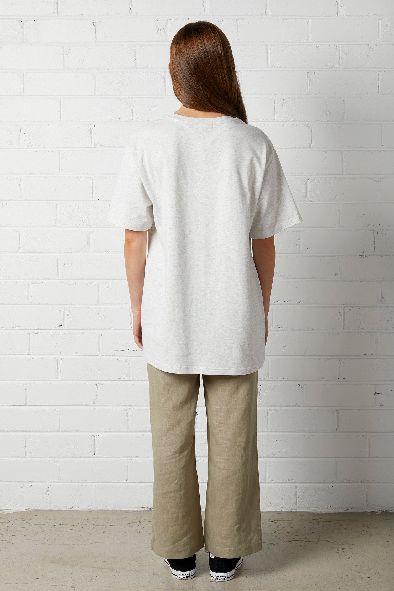Woman with long brown hair wearing Lucy Oversized Tee | NorthByOne in Marle Grey standing against a white brick wall