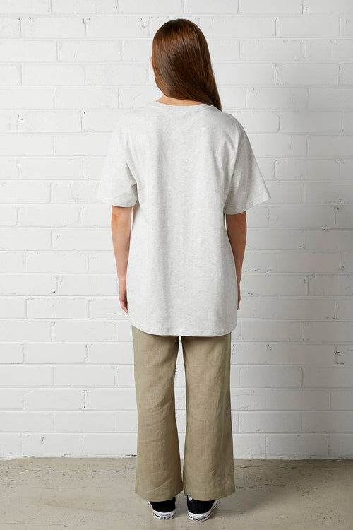 Woman with long brown hair wearing Lucy Oversized Tee | NorthByOne in Marle Grey standing against a white brick wall