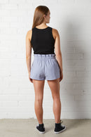 Woman wearing the NorthByOne Aria Paperbag Short, in Indigo and black tank top standing against a white brick wall background