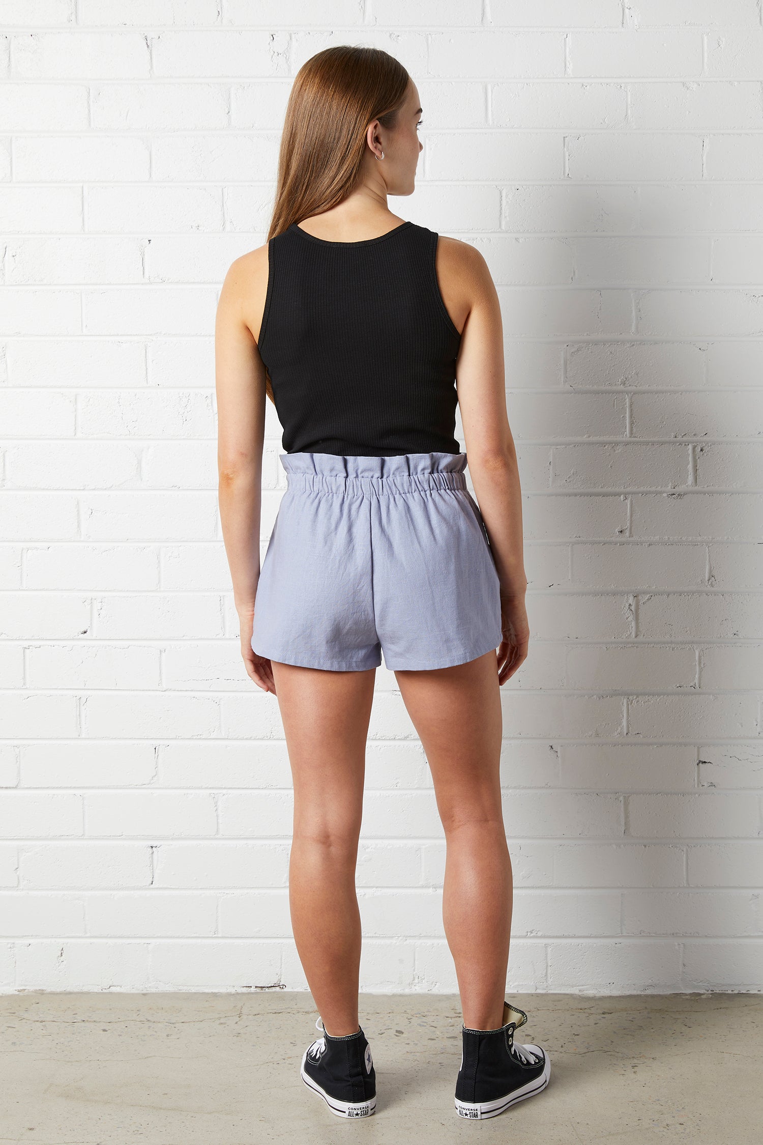 Woman wearing the NorthByOne Aria Paperbag Short, in Indigo and black tank top standing against a white brick wall background
