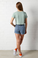 Woman wearing a green cropped t-shirt and NorthByOne Charlotte Denim Short standing with her back against a white brick wall