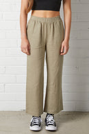 Front view of a person wearing NorthByOne Sadie Linen Pant in Sage Green with an elasticated waistband and casual sneakers against a white brick wall