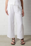 Woman wearing NorthByOne Sadie Linen Pant in white with relaxed fit and elasticated waistband standing against a white brick wall