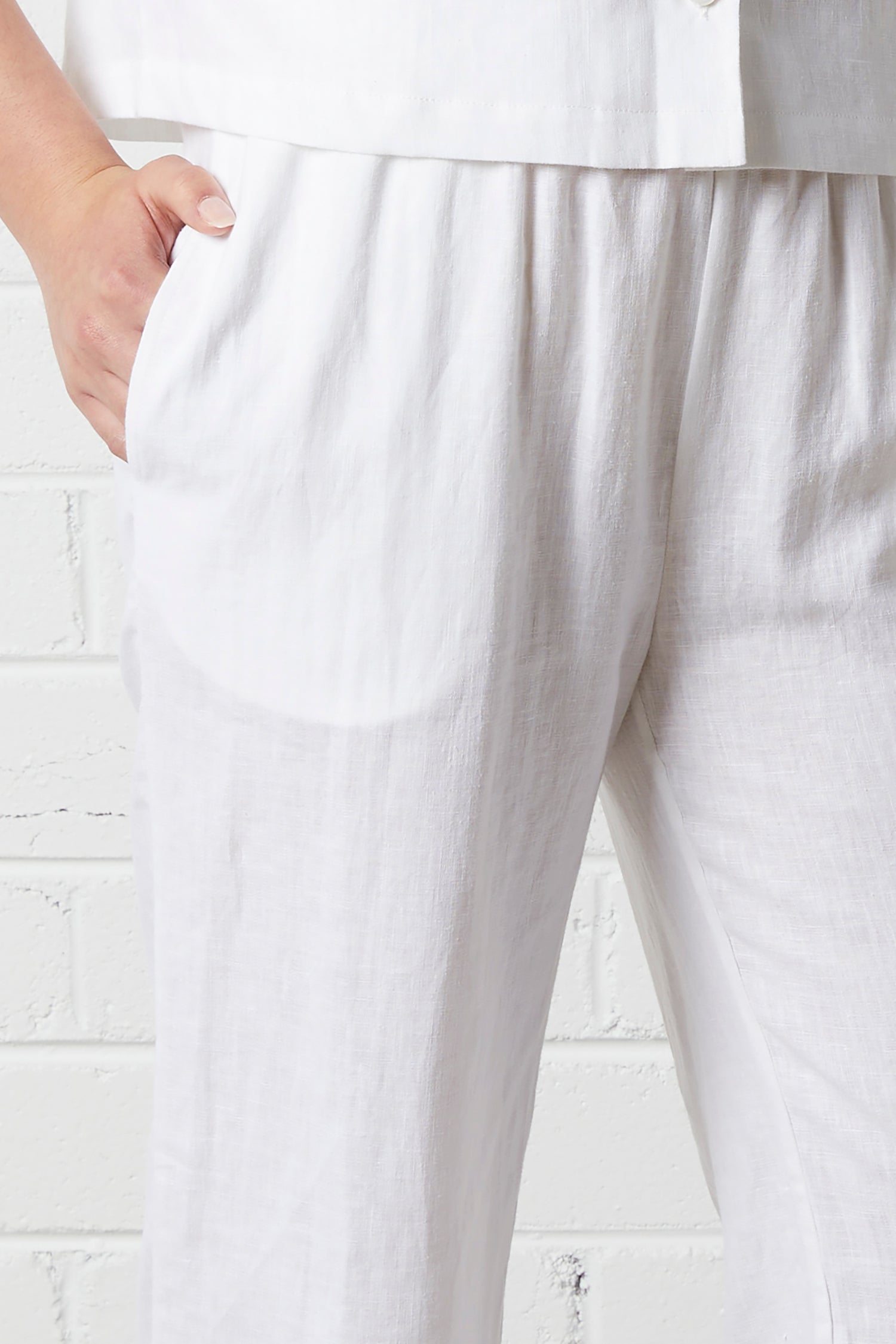 Close-up of person wearing NorthByOne Sadie Linen Pant in white with elasticated waistband and side pockets against a white brick wall