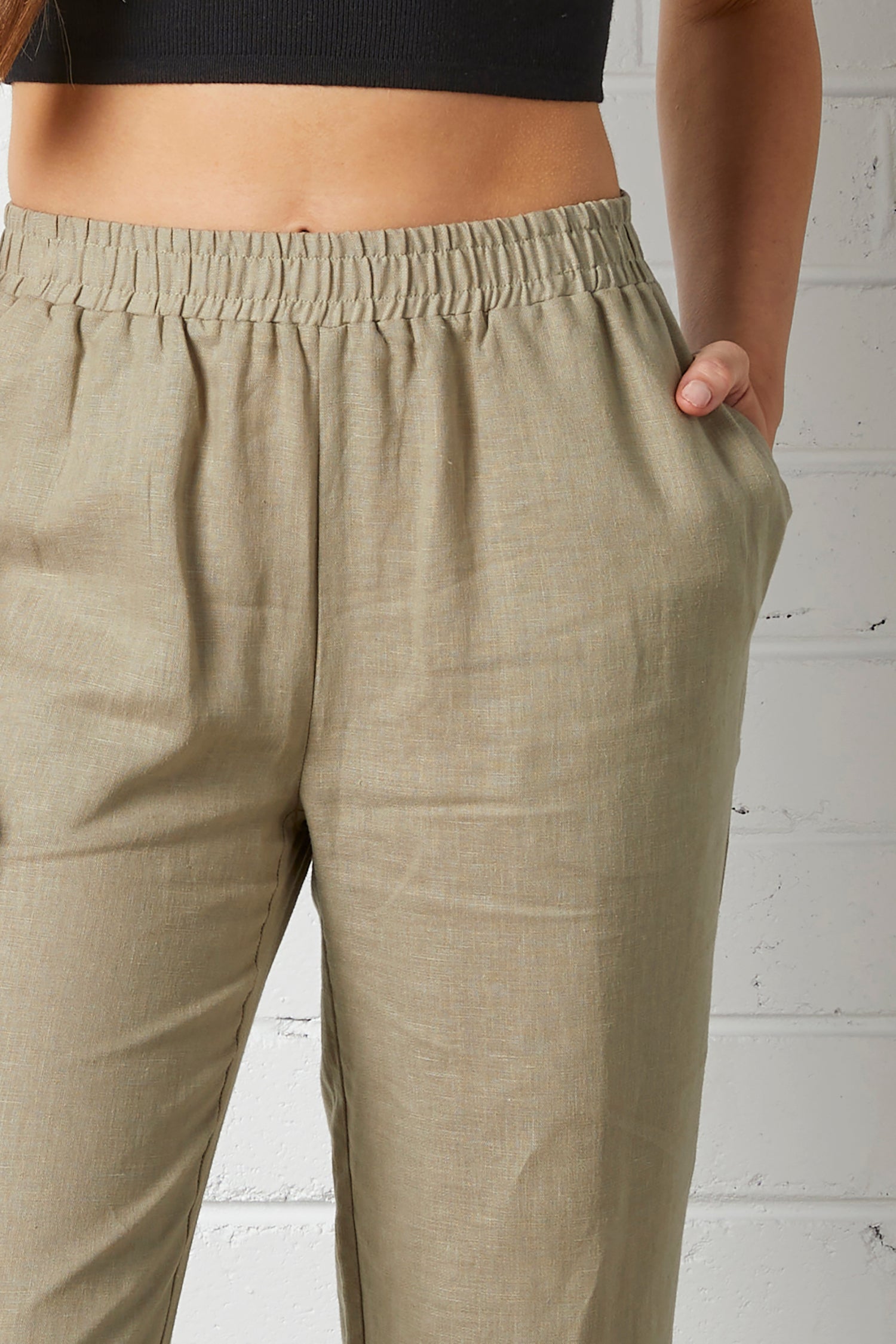 Close-up of a woman wearing NorthByOne Sadie Linen Pant in Sage Green with elastic waistband and side pockets against a white brick wall