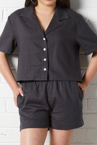 Woman wearing a NorthByOne Zoe Linen Shirt in Charcoal with short sleeves and button front standing against white brick wall