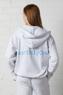 Woman wearing a NorthByOne Olive Zipper Hoodie in Marle Grey with hood up standing against a white brick wall seen from behind