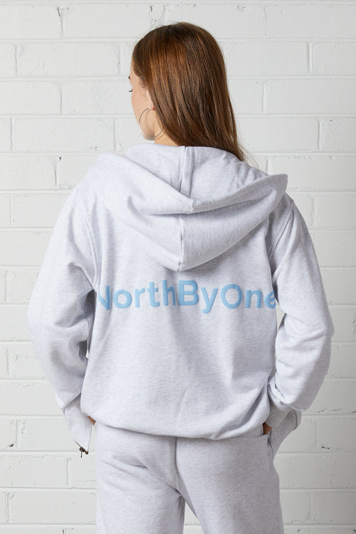 Woman wearing a NorthByOne Olive Zipper Hoodie in Marle Grey with hood up standing against a white brick wall seen from behind