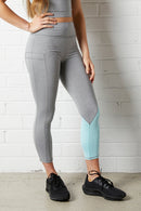 Woman wearing the NorthByOne Cara Three Quarter Tight - Marle Grey / Aqua with black sneakers standing against a white brick wall