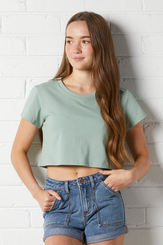 Young woman wearing the NorthByOne Daisy Crop Tee in Sage Green, paired with high waisted denim shorts against a white brick wall