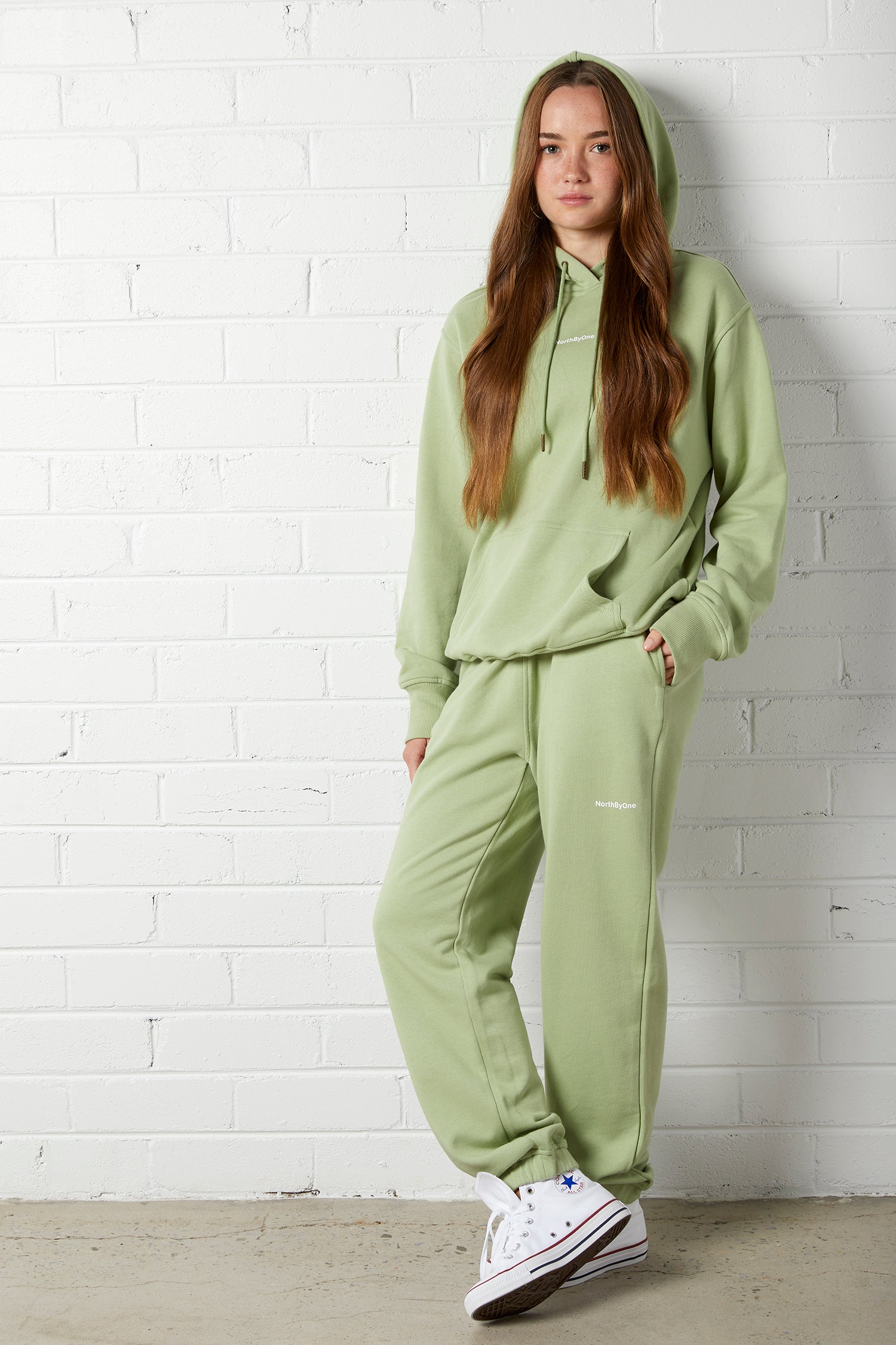 Young woman wearing the NorthByOne Emma Baggy Sweat Pant in Avocado, with matching hoodie and white sneakers against white brick wall
