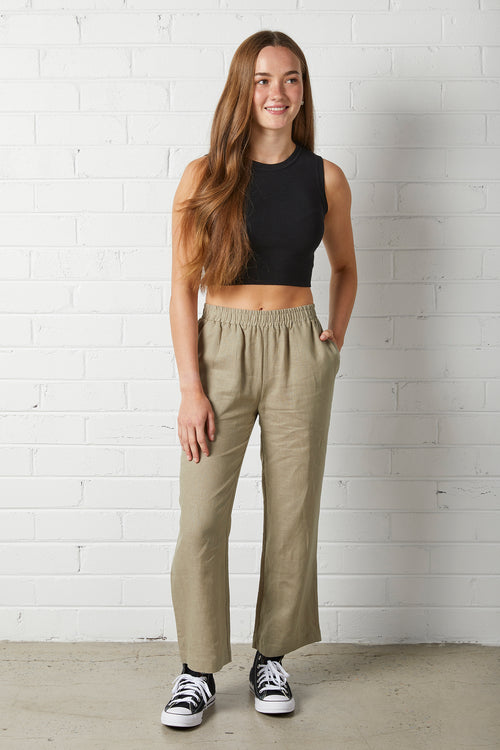 Young woman wearing NorthByOne Sadie Linen Pant in Sage Green and a black sleeveless crop top standing against a white brick wall