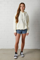 Young woman wearing a NorthByOne Frankie Hoodie in vanilla with denim shorts and black sneakers against a white brick wall