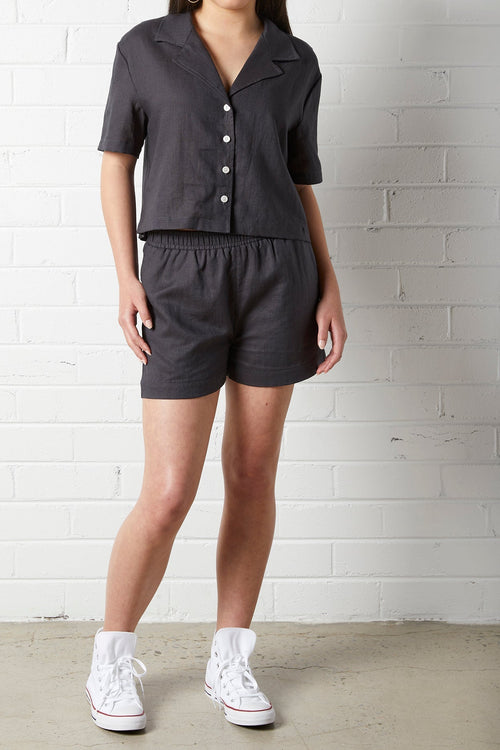 Woman wearing a NorthByOne Zoe Linen Shirt in Charcoal with matching shorts and white high-top sneakers against a white brick wall