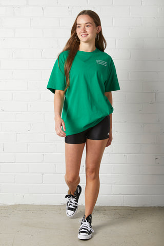 Woman wearing Lucy Oversized Tee | NorthByOne in Emerald with black shorts and black sneakers standing against white brick wall