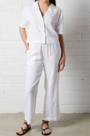 Woman wearing NorthByOne Zoe Linen Shirt in White with matching white linen pants and black sandals against a white brick wall